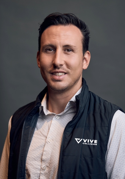 A man with short dark hair is smiling slightly while wearing a light-patterned button-down shirt and a black VIVE Collision branded vest, posed against a neutral gray background.